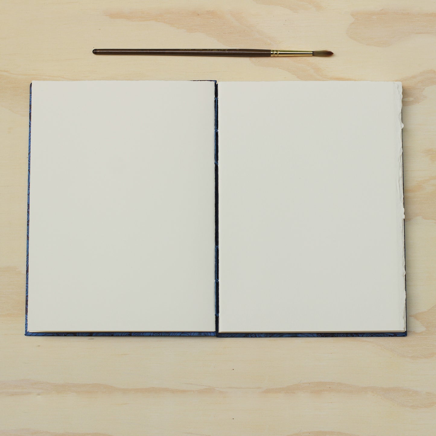 An opened blank art journal or sketchbook with blank white pages, placed on a light wood surface with a small paintbrush above it.