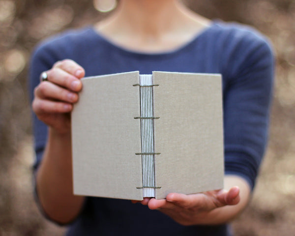 Load image into Gallery viewer, Hands holding square sketchbook in bisque with olive green binding thread.
