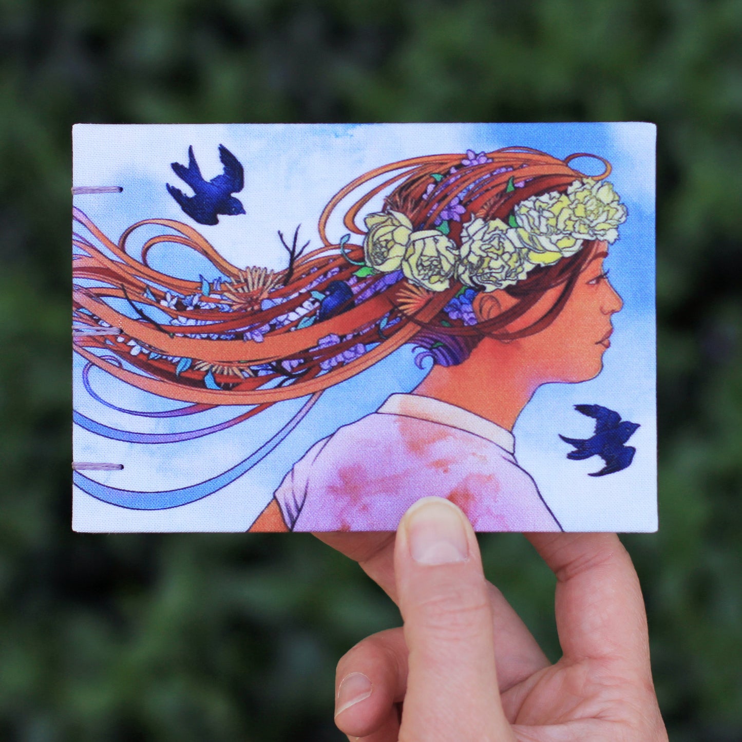 hand holding Emmylou Smith Lake Michigan Book Press sketchbook. Leafy green background.