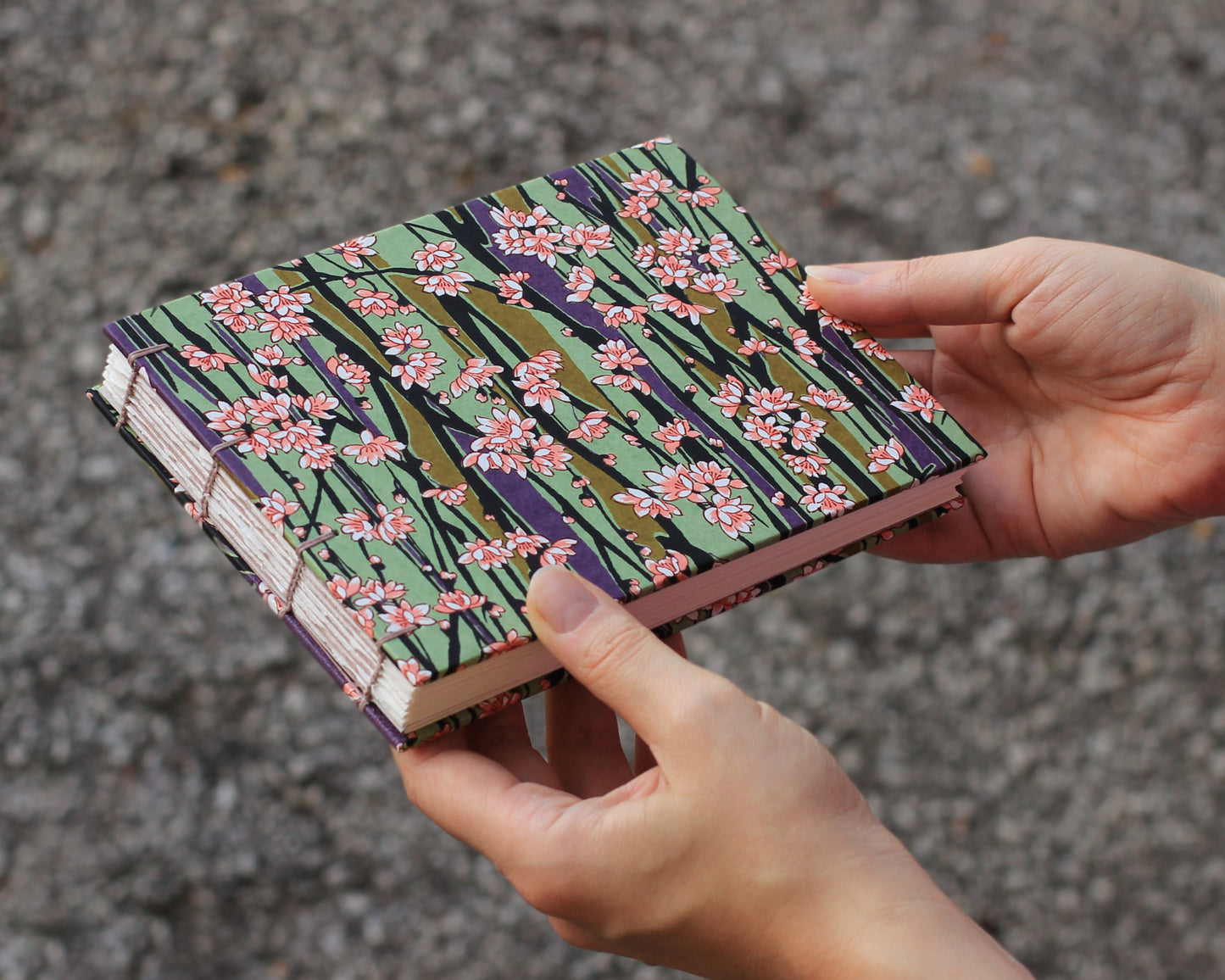 Hand holding a sketchbook with a floral pattern on the cover, photographed against gravel background.