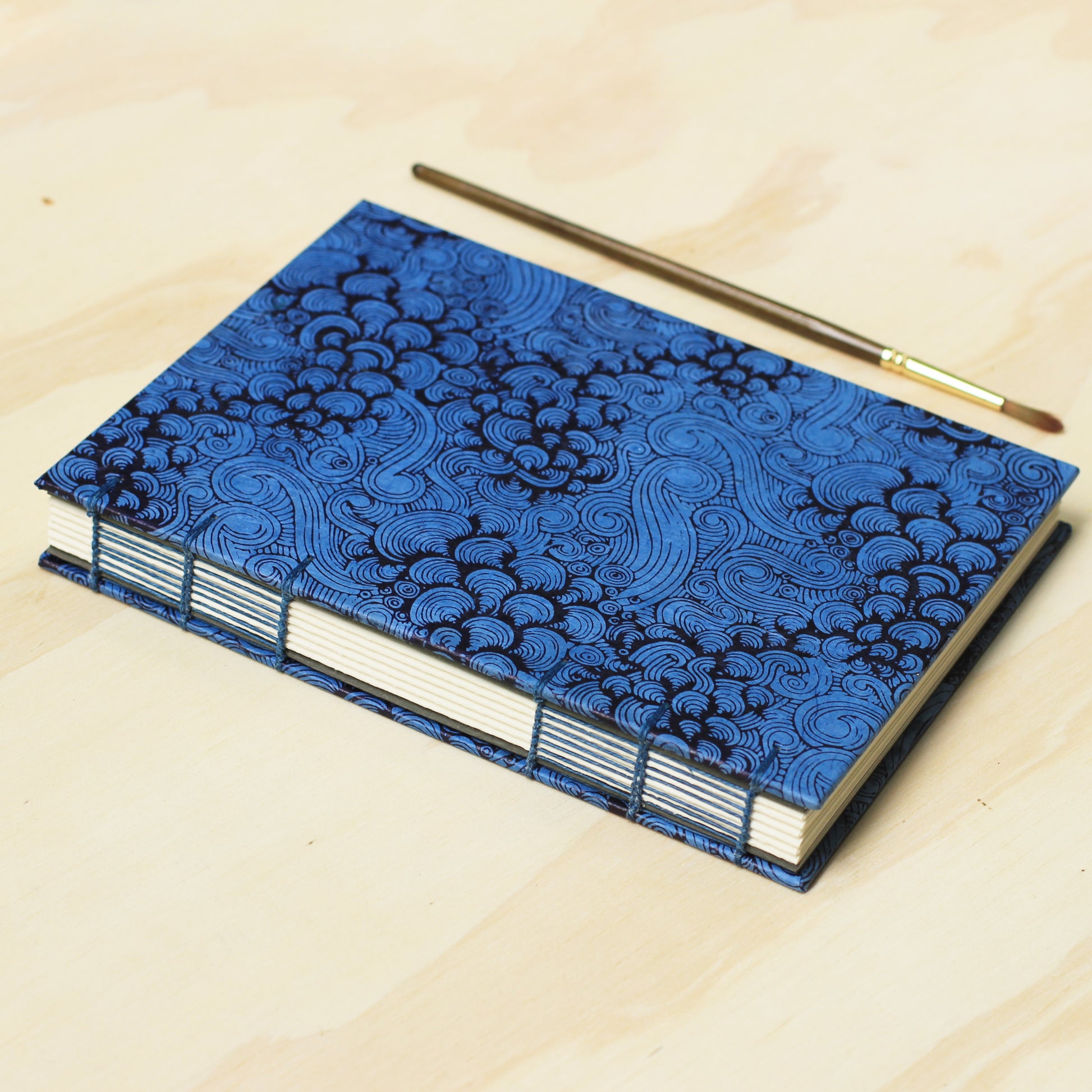 A handmade blue and black patterned journal with white page edges, shown on a light wood background with a small paintbrush resting beside it.