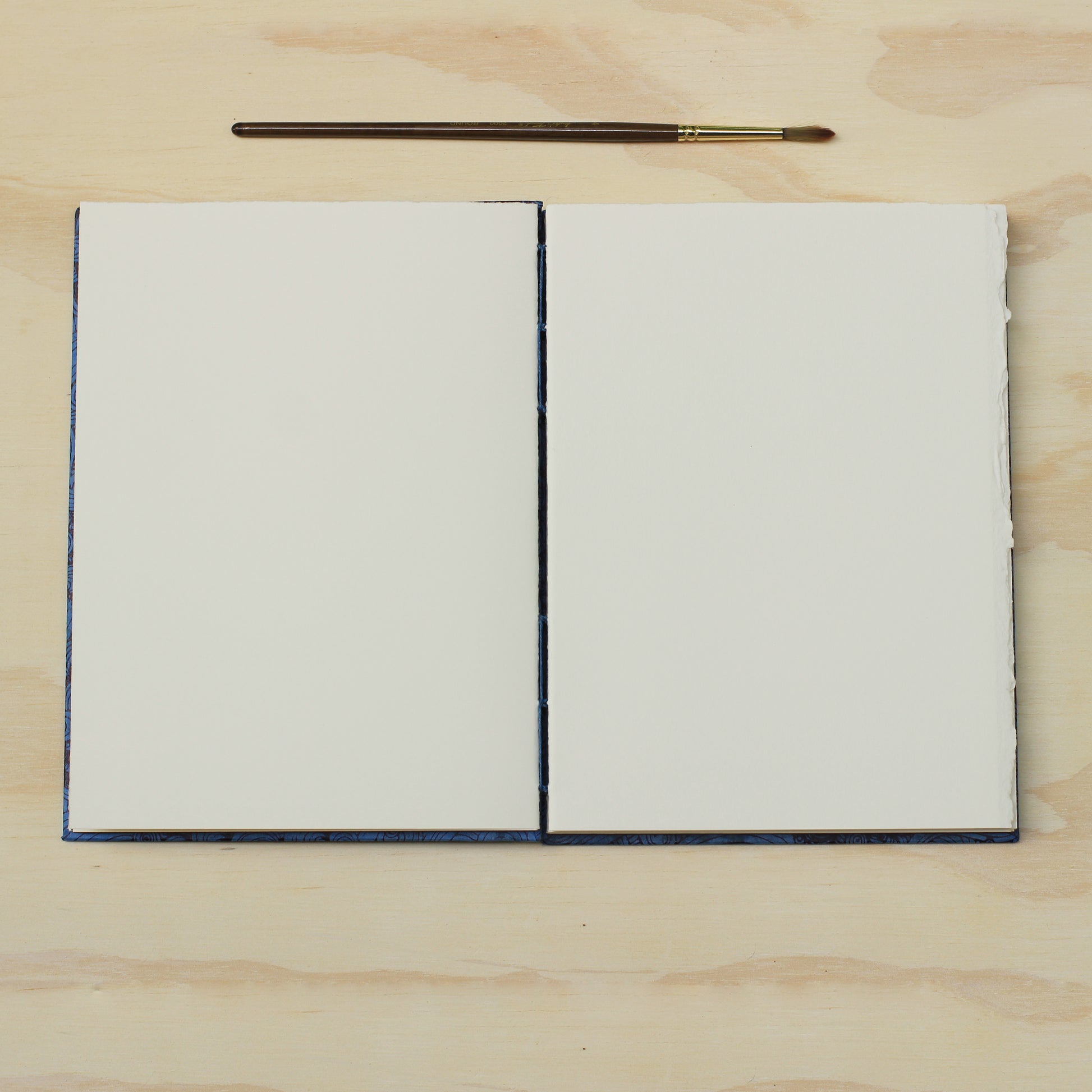 An opened blank art journal or sketchbook with blank white pages, placed on a light wood surface with a small paintbrush above it.