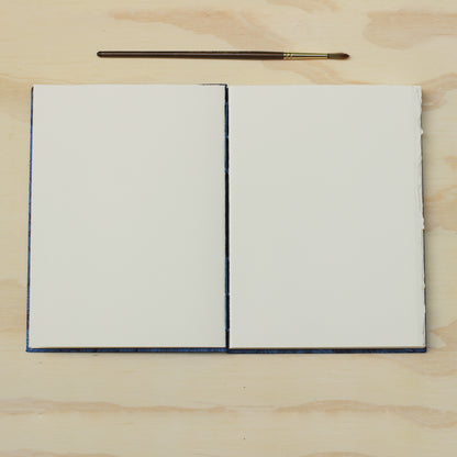 An opened blank art journal or sketchbook with blank white pages, placed on a light wood surface with a small paintbrush above it.