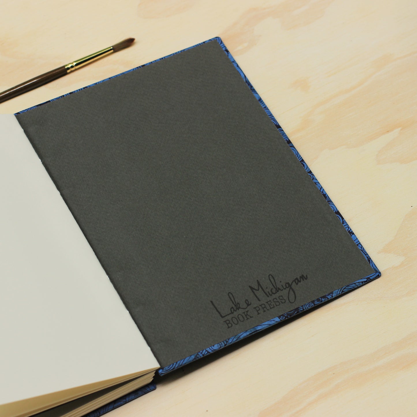 A dark gray hardcover book with a blue patterned edge and the embossed text 'Lake Michigan Book Press' on the cover, photographed open on a light wood surface with a paintbrush in the corner.