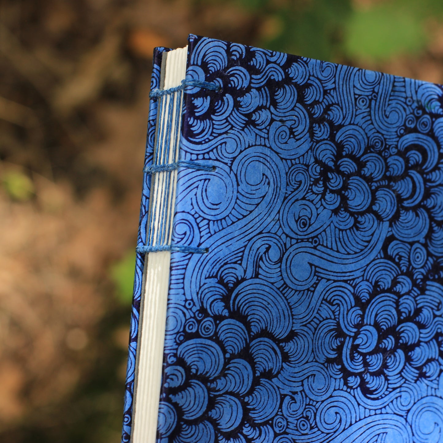 A close-up of a handmade journal with a blue and black spiral patterned cover, bound with blue thread, set against a blurred outdoor background.