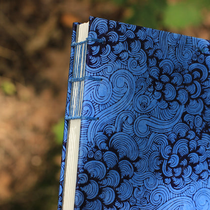 A close-up of a handmade journal with a blue and black spiral patterned cover, bound with blue thread, set against a blurred outdoor background.