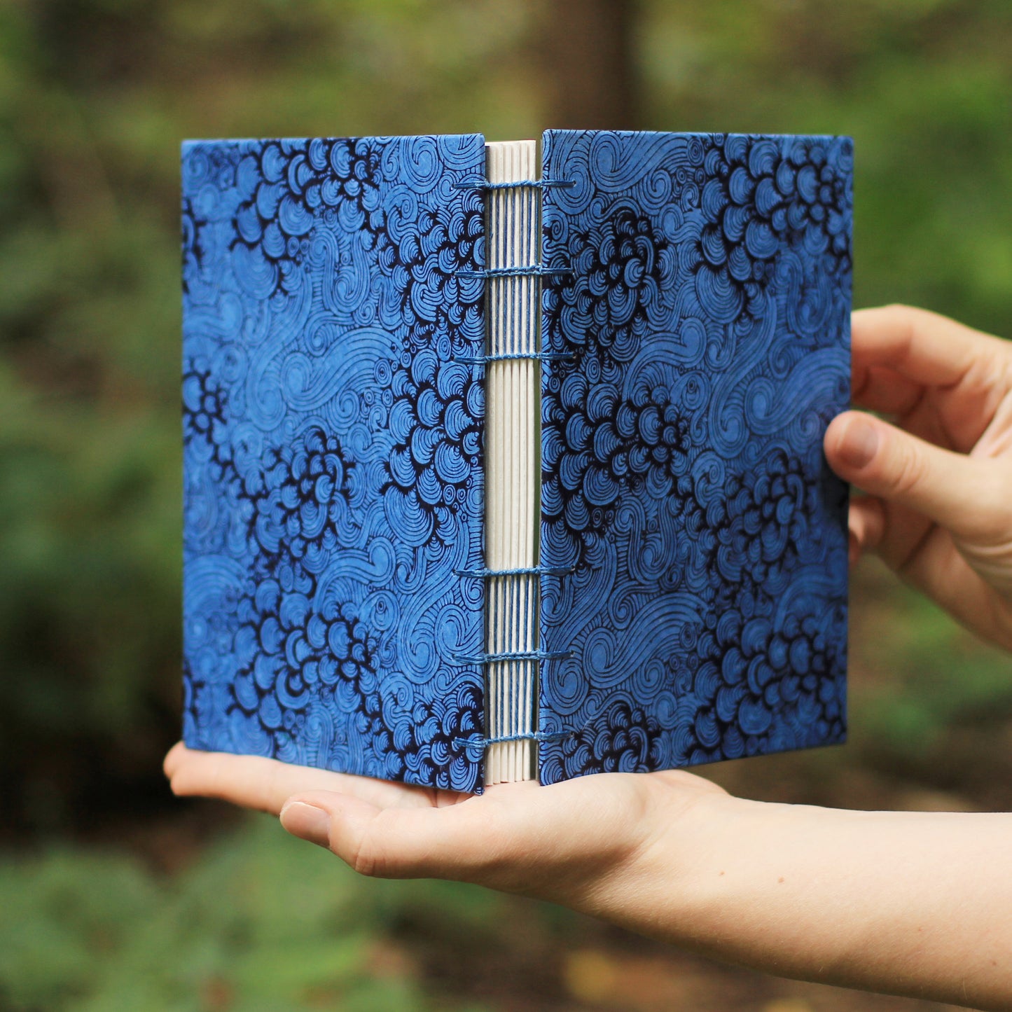 Hands holding a blue sketchbook with a black spiral pattern cover, set against a blurred green outdoor background.