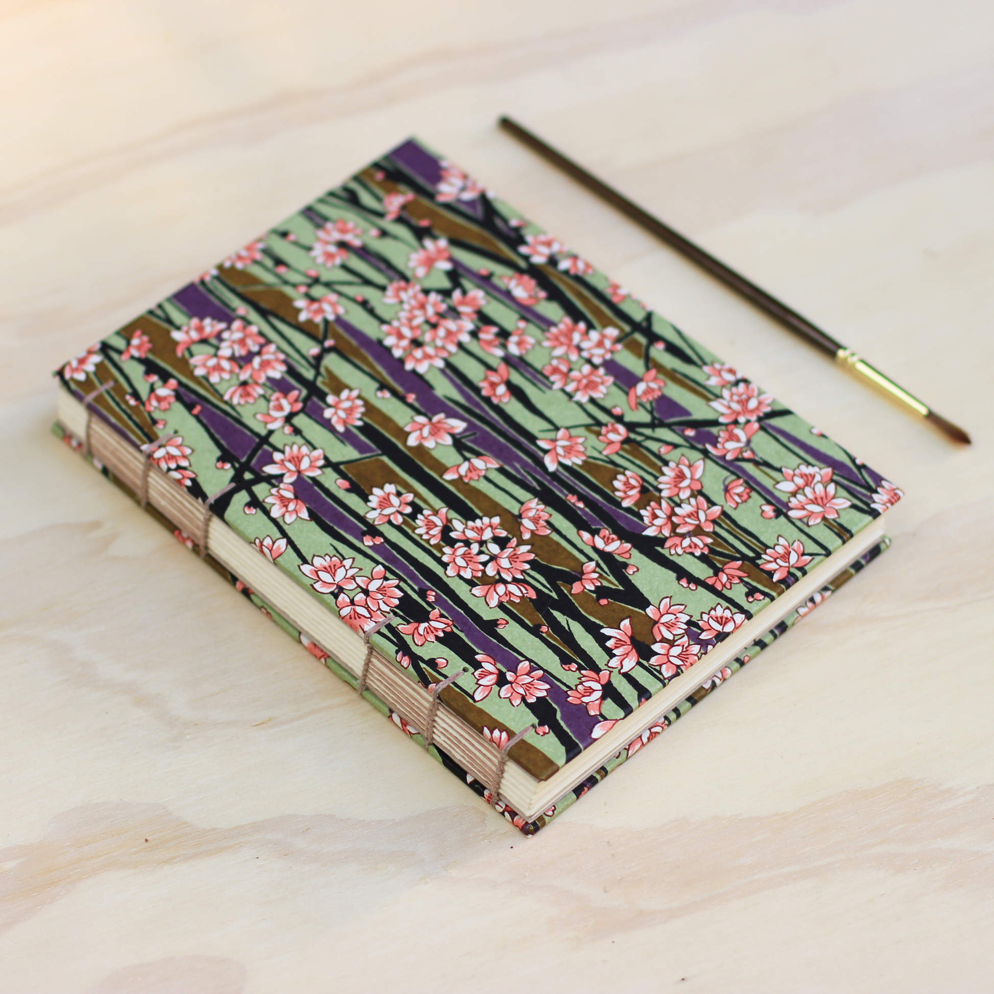 A handmade Japanese-style paper notebook with a cherry blossom pattern, shown on a light wood surface next to a paintbrush.