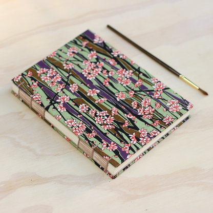 A handmade Japanese-style paper notebook with a cherry blossom pattern, shown on a light wood surface next to a paintbrush.