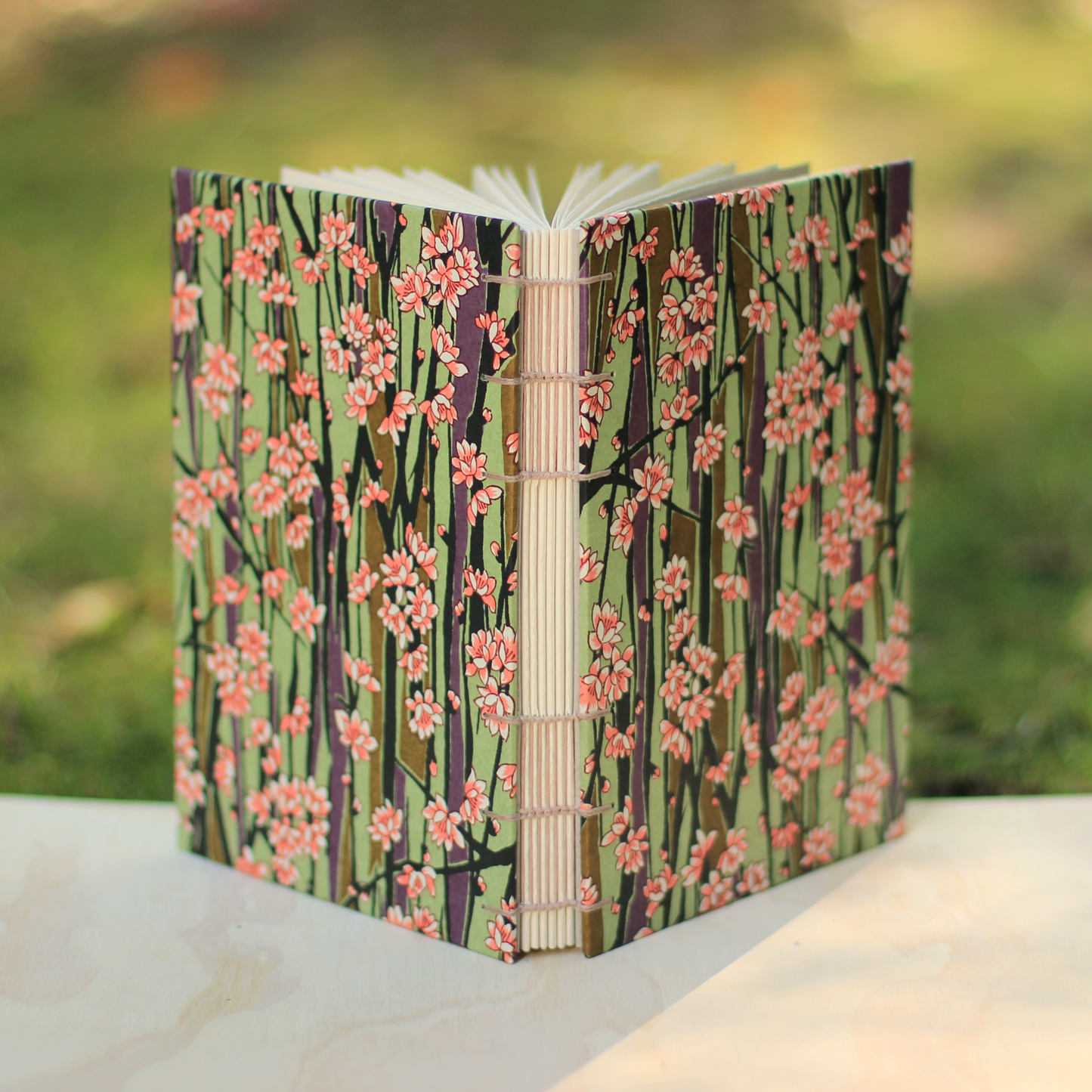 A handmade open journal with a green floral patterned cover, photographed outdoors on a light stone surface with a blurred green background.