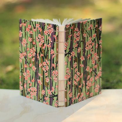 A handmade open journal with a green floral patterned cover, photographed outdoors on a light stone surface with a blurred green background.