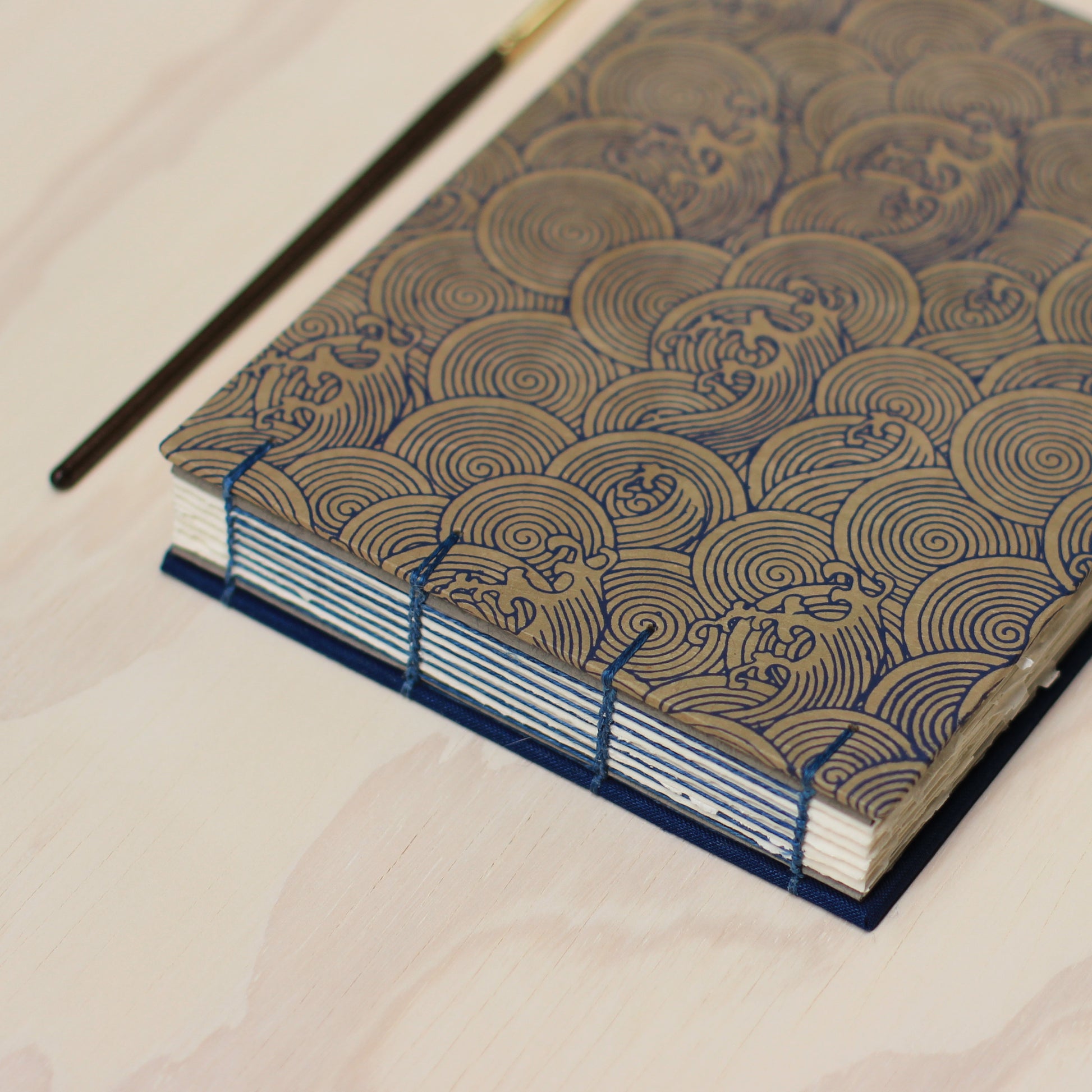 A close-up of a handmade sketchbook with a dark blue fabric back cover and a patterned blue wave and beige front cover, photographed on a light wood surface.