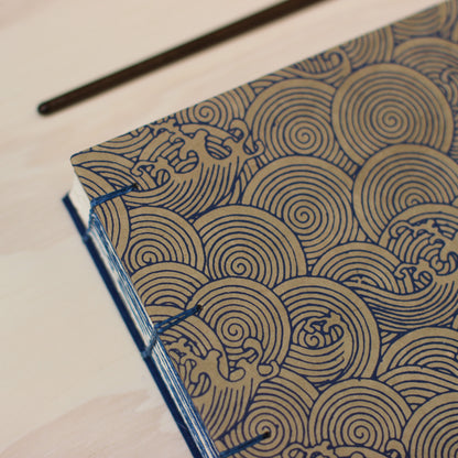 A close-up of a handmade sketchbook with a tan cover featuring a navy blue wave pattern, bound with blue thread, on a light wood background.