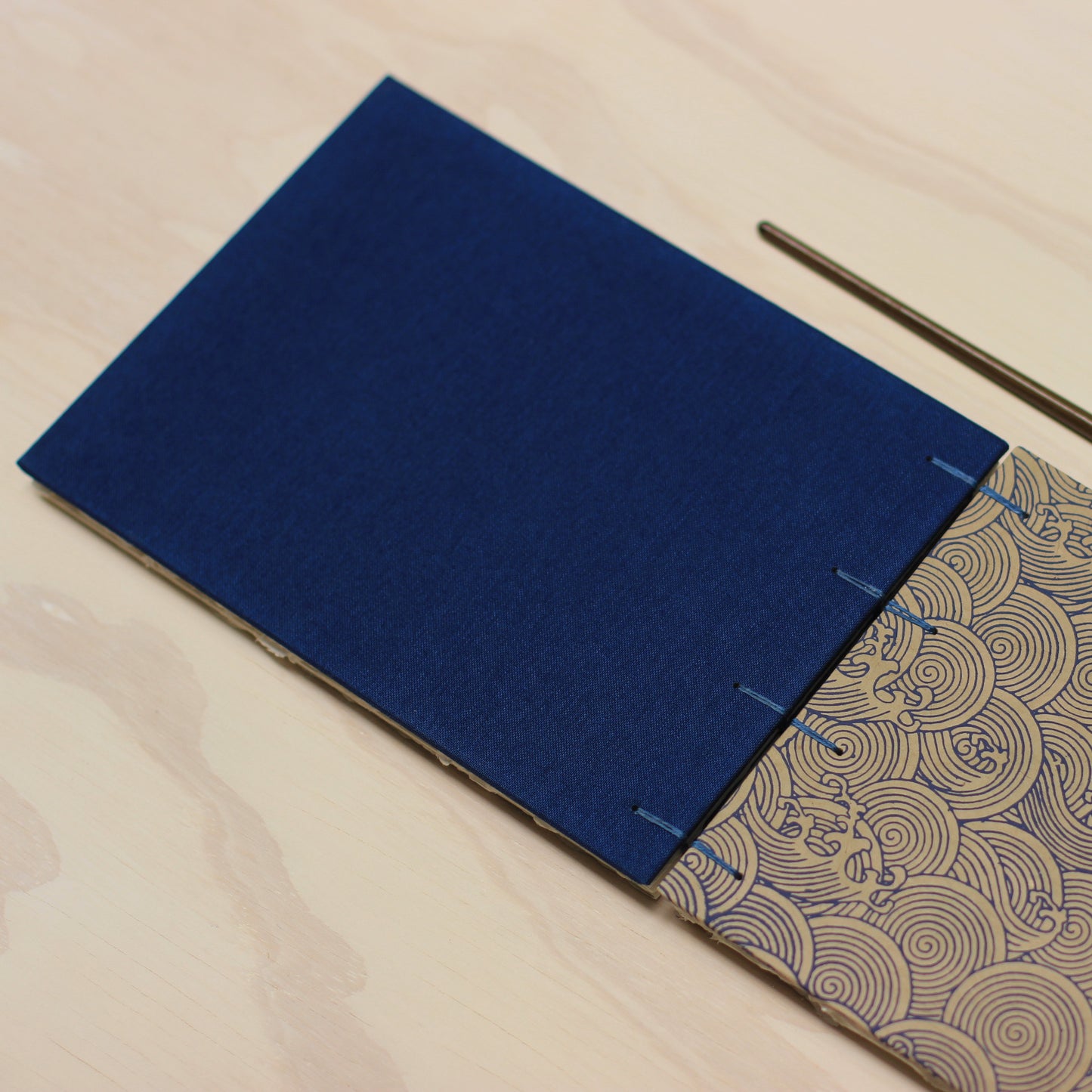 A close-up of a handmade sketchbook with a dark blue fabric back cover and a patterned blue wave and beige front cover, photographed on a light wood surface.