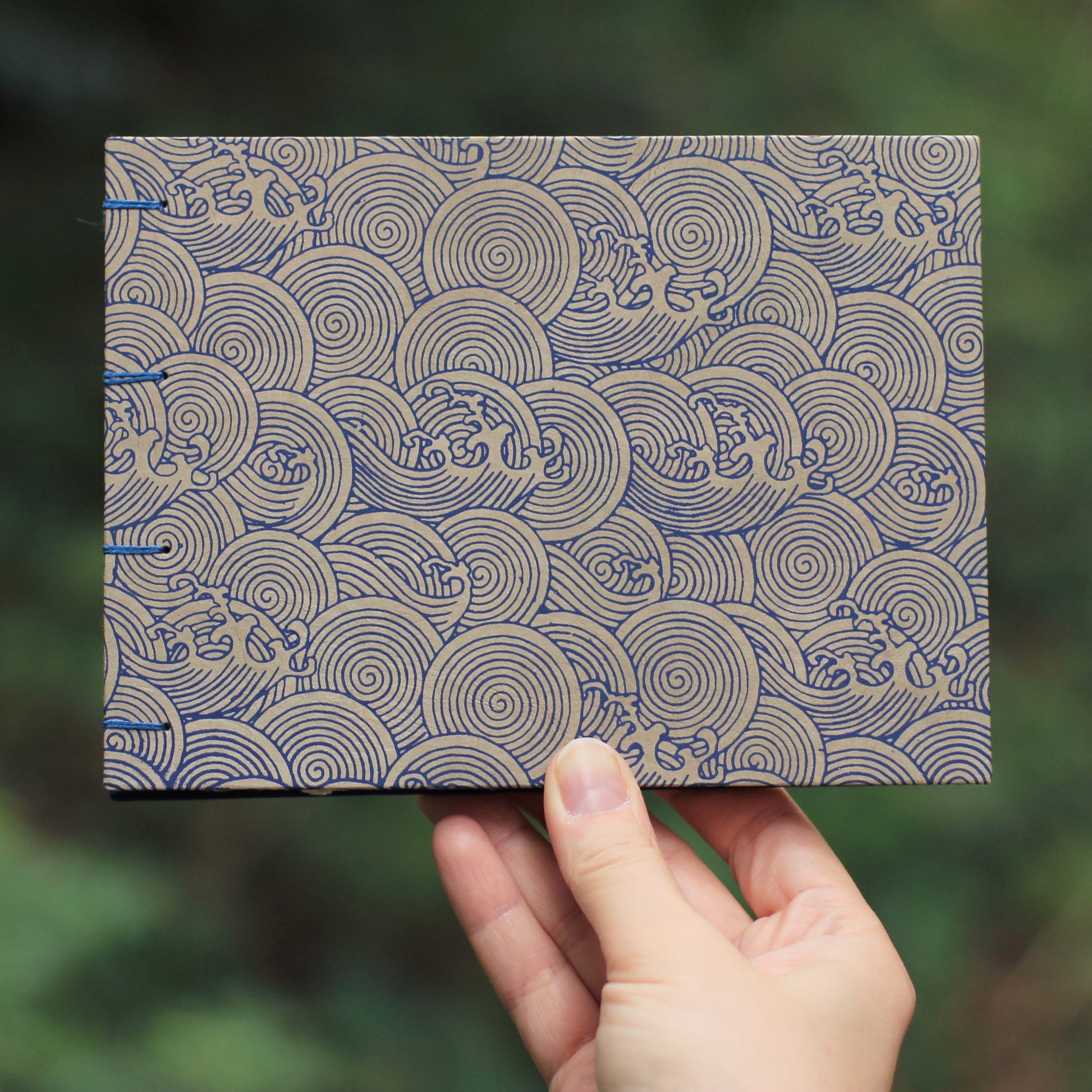 A hand holding a handmade sketchbook featuring a blue and beige wave pattern against a blurred green outdoor background.