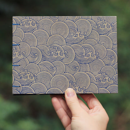 A hand holding a handmade sketchbook featuring a blue and beige wave pattern against a blurred green outdoor background.