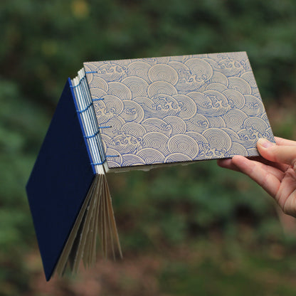 A hand holding a handmade sketchbook with a blue patterned cover and visible page edges, set against a blurred green outdoor background.