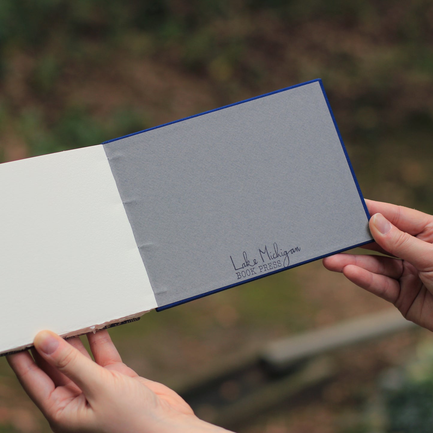A person holds an open sketchbook outdoors. The right side is a textured gray interior back cover with the printed text 'Lake Michigan Book Press' in black script, and the left side is white paper.