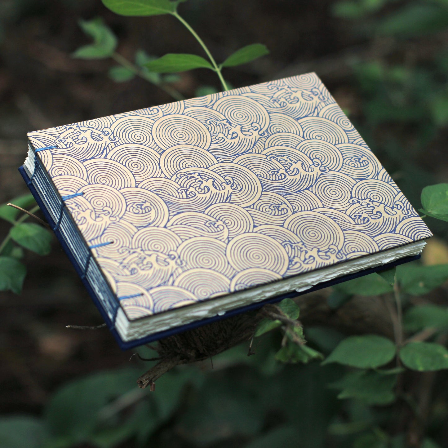 A handmade sketchbook with a blue and beige wave pattern cover, held up by a branch in a natural outdoor setting with green leaves.