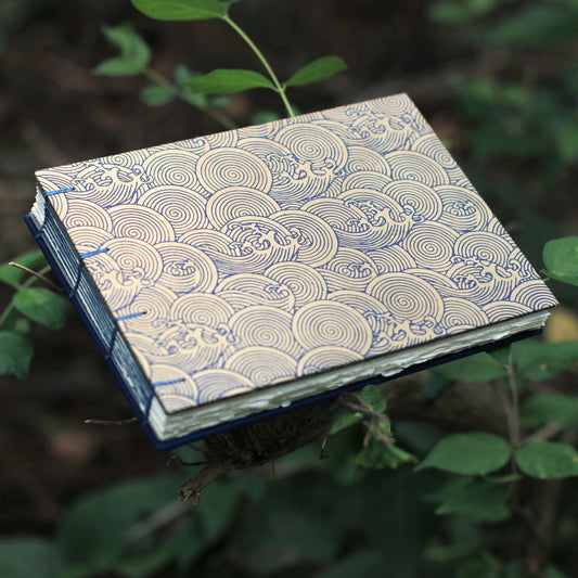 A handmade sketchbook with a blue and beige wave pattern cover, held up by a branch in a natural outdoor setting with green leaves.