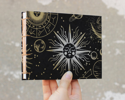 black sketchbook with silver sun on cover held by hand on gray background