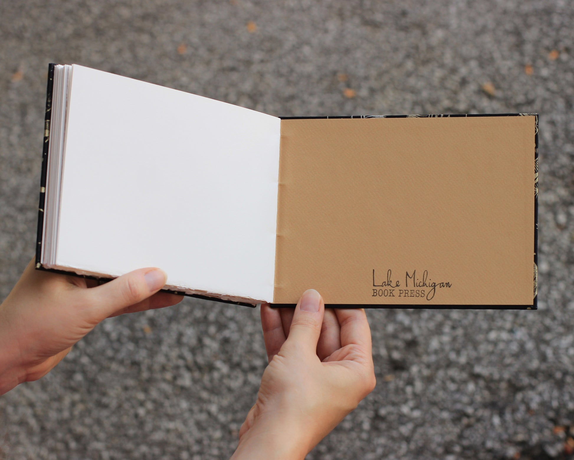 Blank sketchbook open to back interior cover, held open by two hands. Back cover reads "lake michigan book press" in script on a tan background.