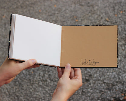 Blank sketchbook open to back interior cover, held open by two hands. Back cover reads "lake michigan book press" in script on a tan background.