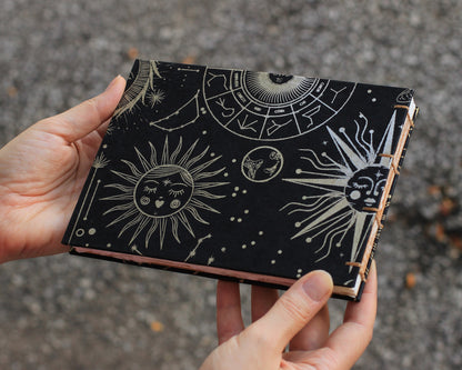 black sketchbook with gold suns with faces sitting held by two hands against gravel background
